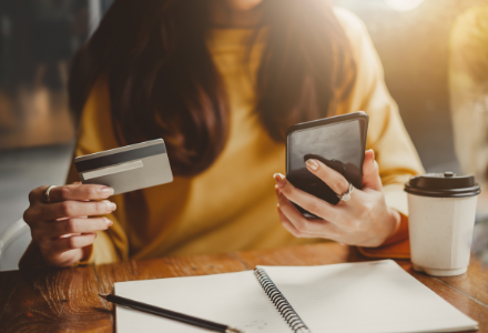 Lady holding a mobile phone and credit card, next to her is a coffee, pen and paper.